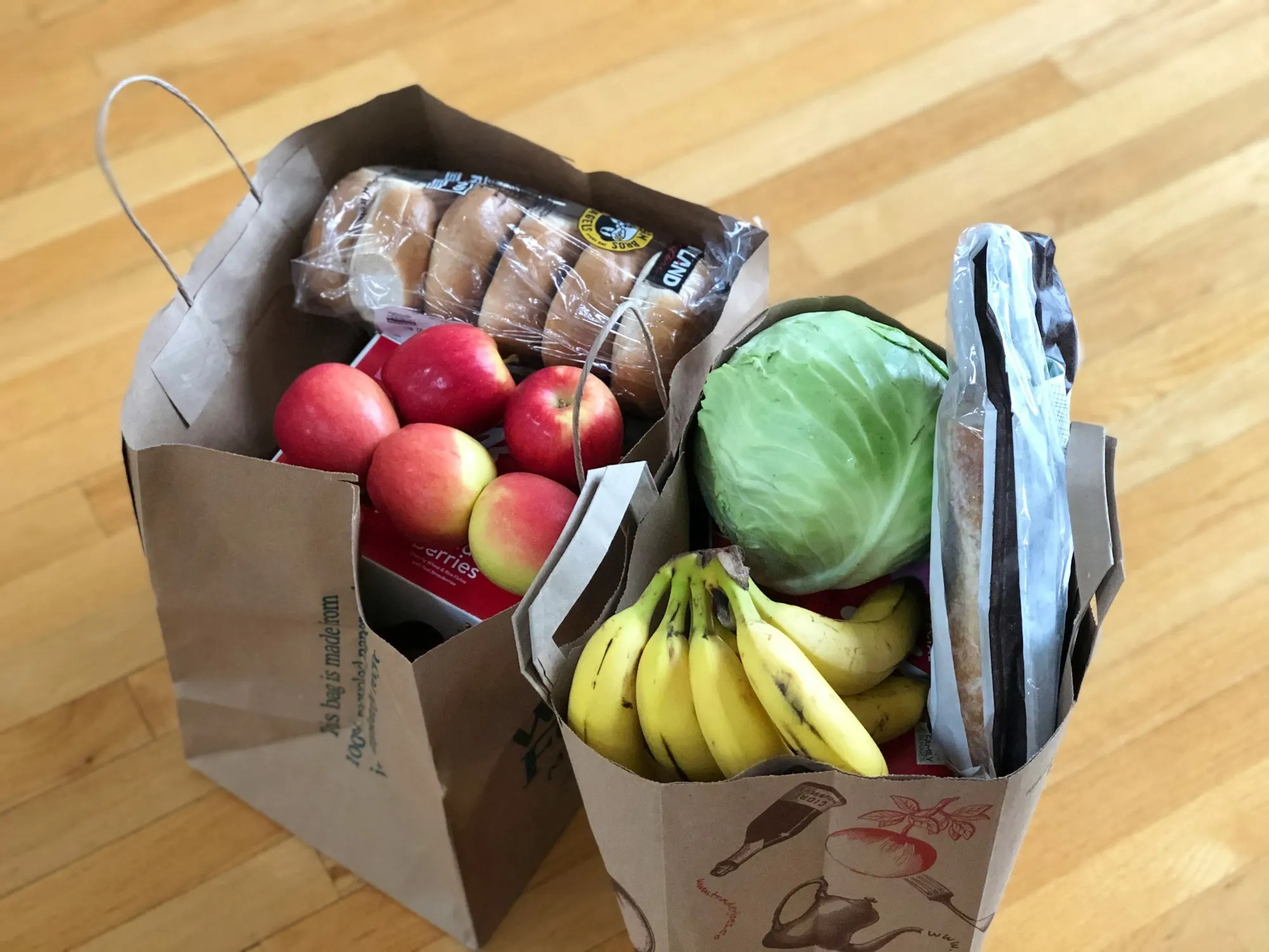 Two paper grocery bags holding apples, bananas, a cabbage and bread, seen from above on a wooden floor
